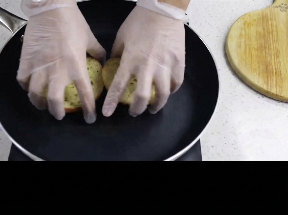 Gloved hands placing the buttered side of burger buns face down into a hot black frying pan.