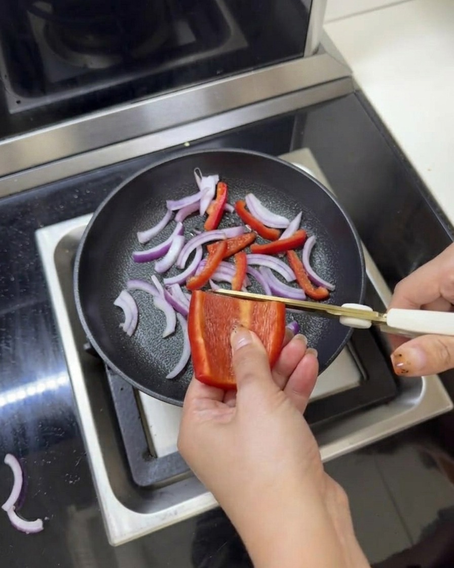 Sliced red onions and bell peppers in a frying pan being seasoned with salt and pepper using kitchen scissors.