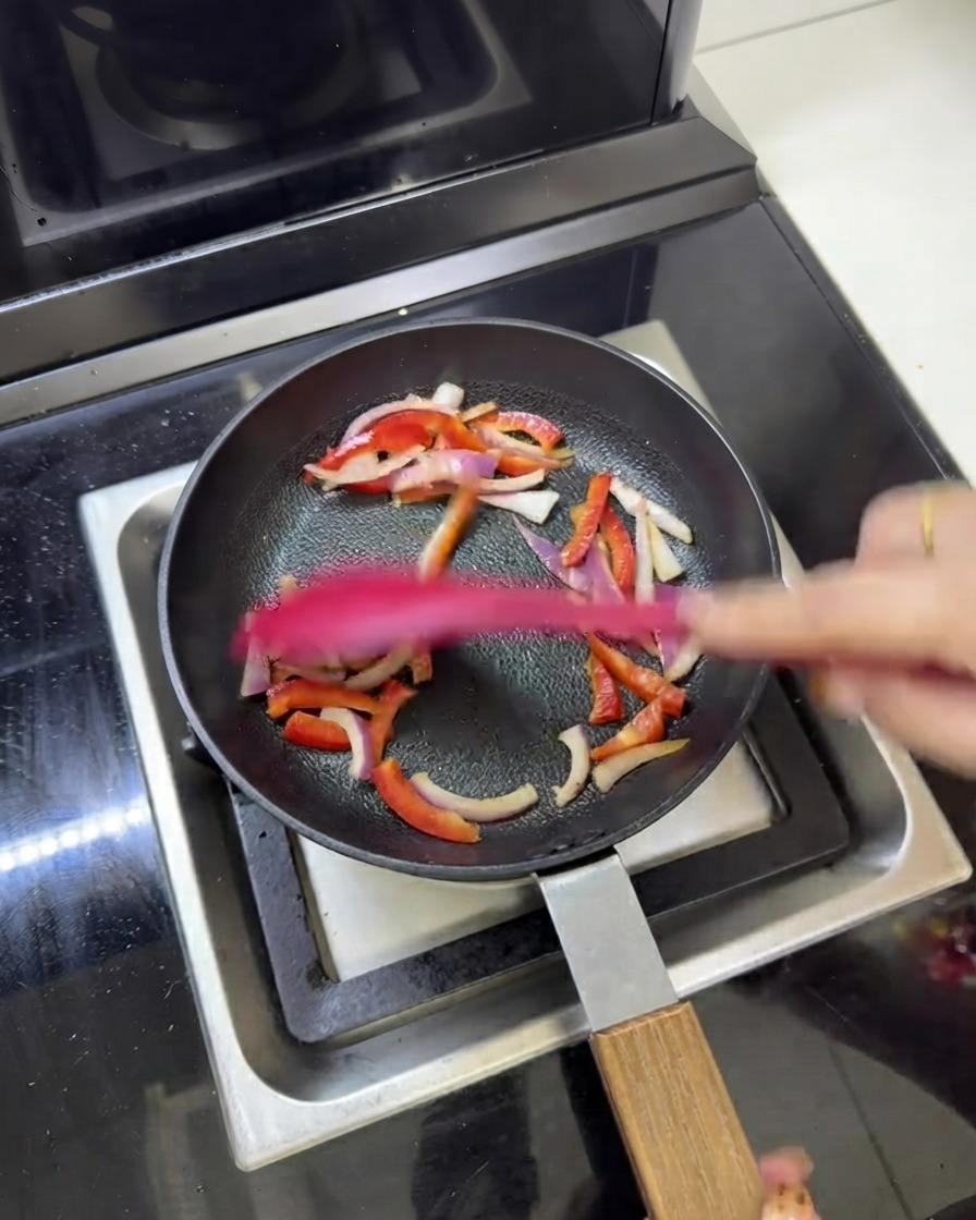 Pre-cooked meat strips being stir-fried alongside red onions and bell peppers in a frying pan.