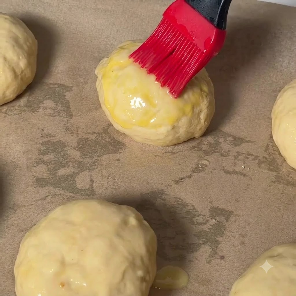A red pastry brush applying egg wash onto the top of a cheeseburger dough ball on baking paper.