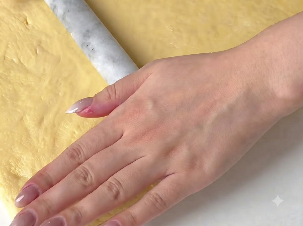 A hand using a marble rolling pin to flatten out a yellow dough sheet on a surface.