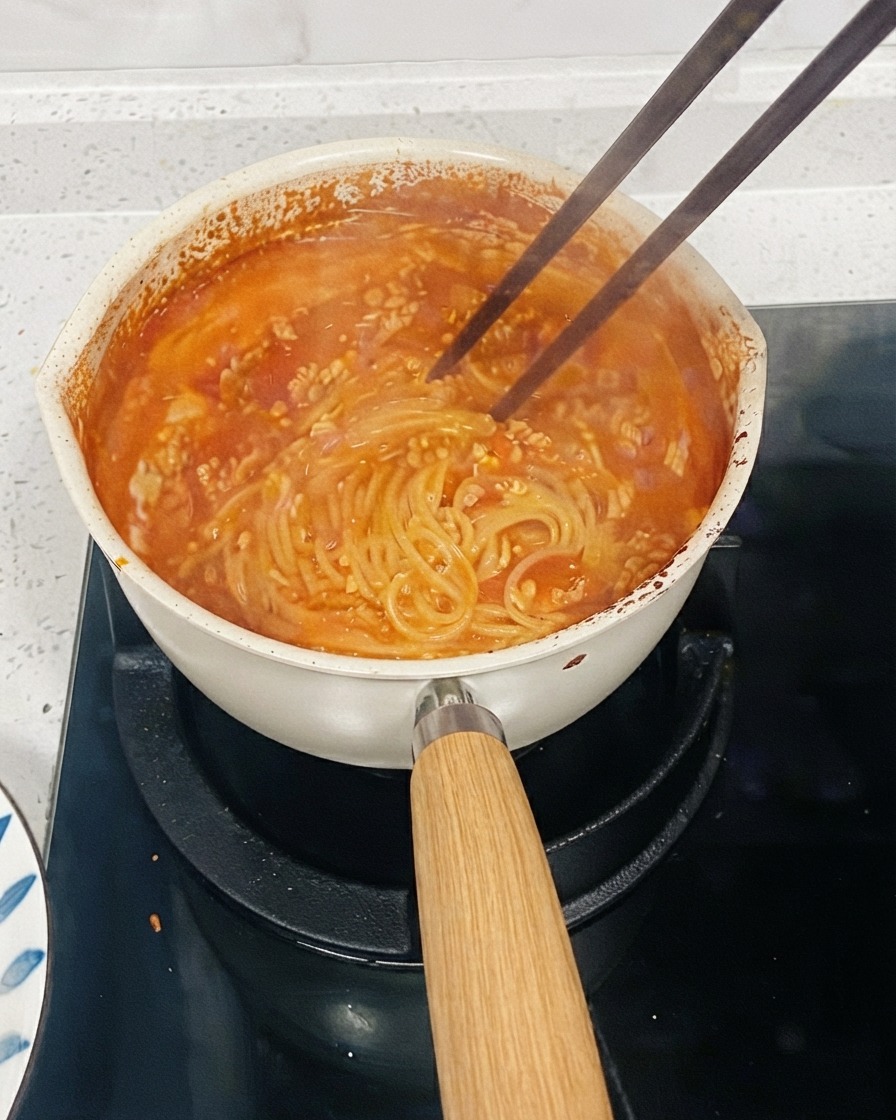 A pot of tomato-based pasta simmering on a stove with chopsticks stirring the noodles to thicken the sauce.