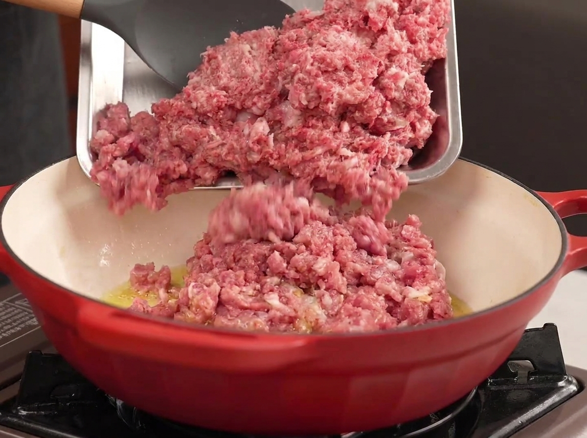 Ground beef and pork being poured from a metal tray into a red pot with hot oil.
