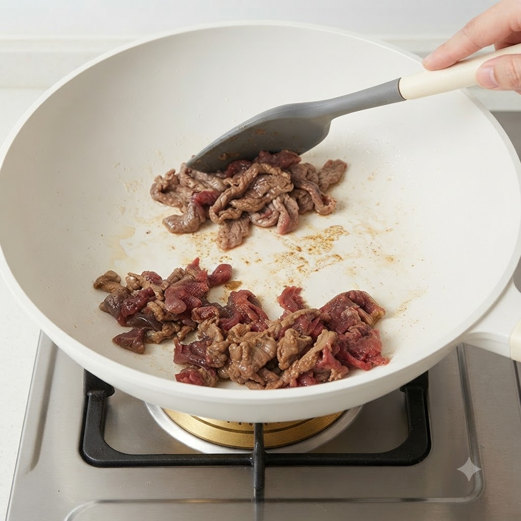 Marinated beef slices being stir-fried in a white pan using a grey silicone spatula.