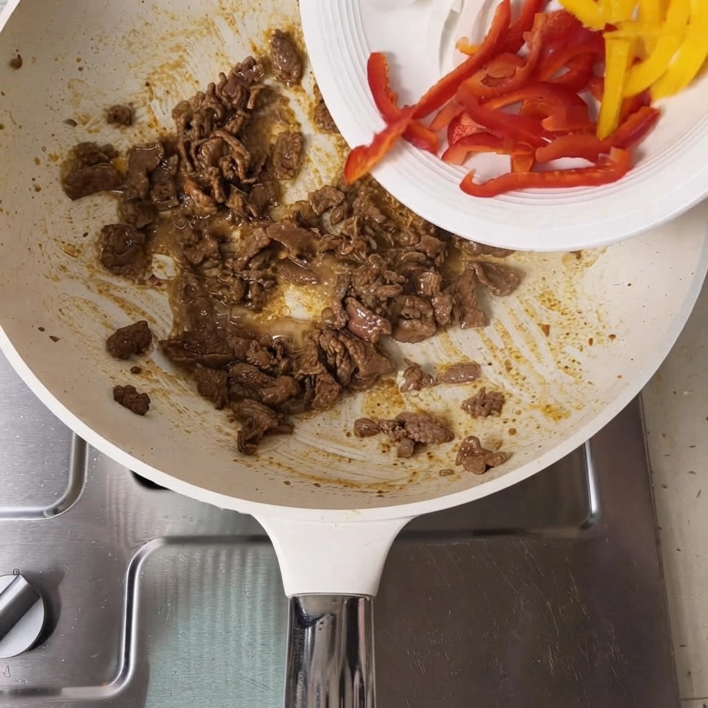 Sliced red and yellow bell peppers being poured from a white plate into a pan containing cooked beef slices.