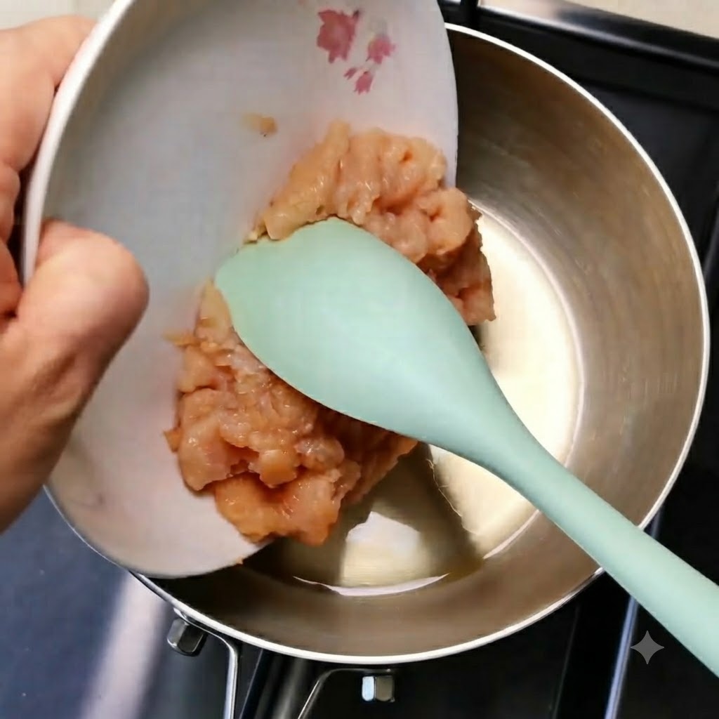 Minced chicken meat sliding from a white plate into a metal pan using a green spatula.