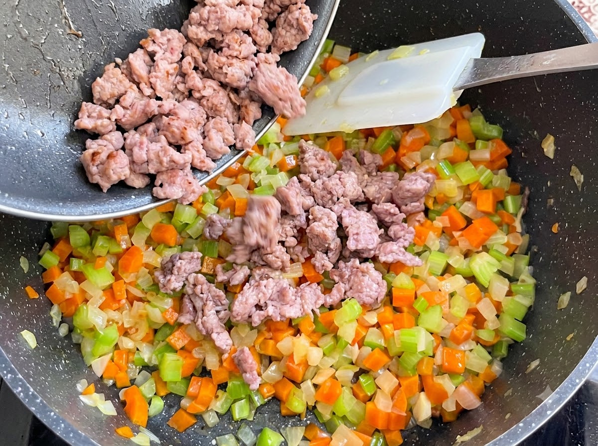 Cooked sausage meat being poured from a pan into the vegetable pot and mixed in.