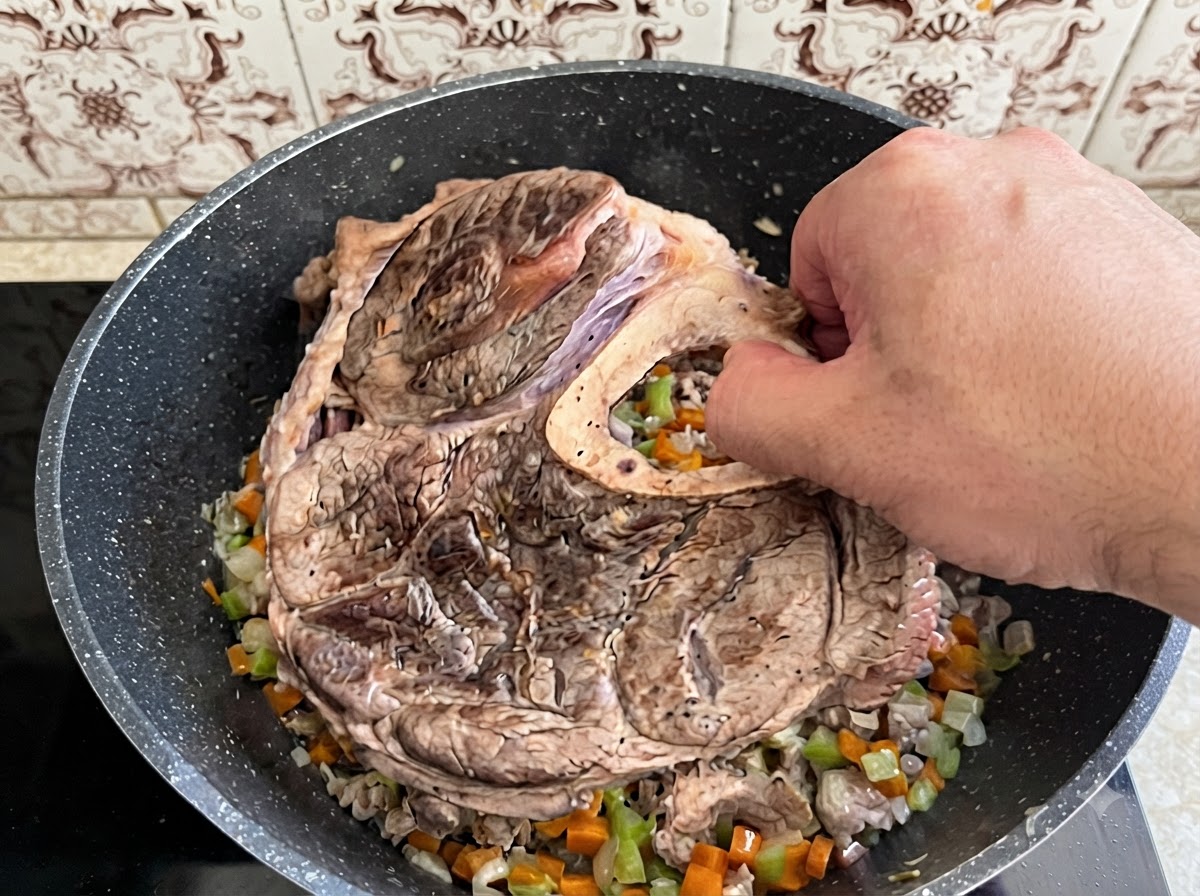 A hand placing a large piece of seared beef shank and bone into a pot filled with cooked diced vegetables.