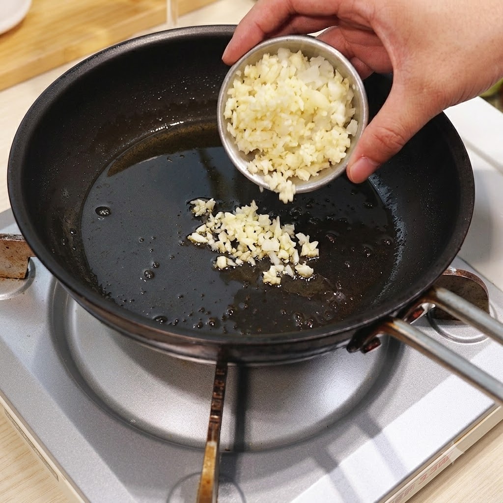 Minced garlic and onion being poured from a small bowl into a hot frying pan with oil.