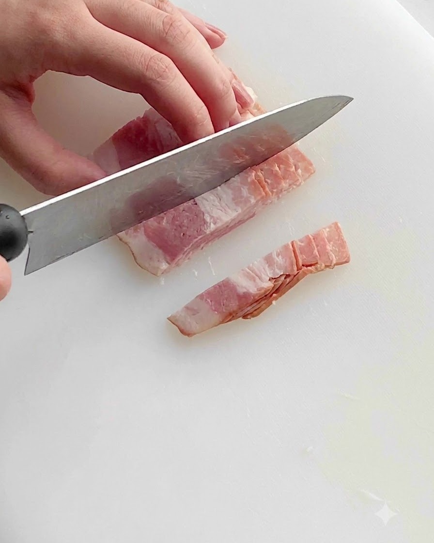 A kitchen knife slicing raw bacon strips into small uniform pieces on a white plastic cutting board.