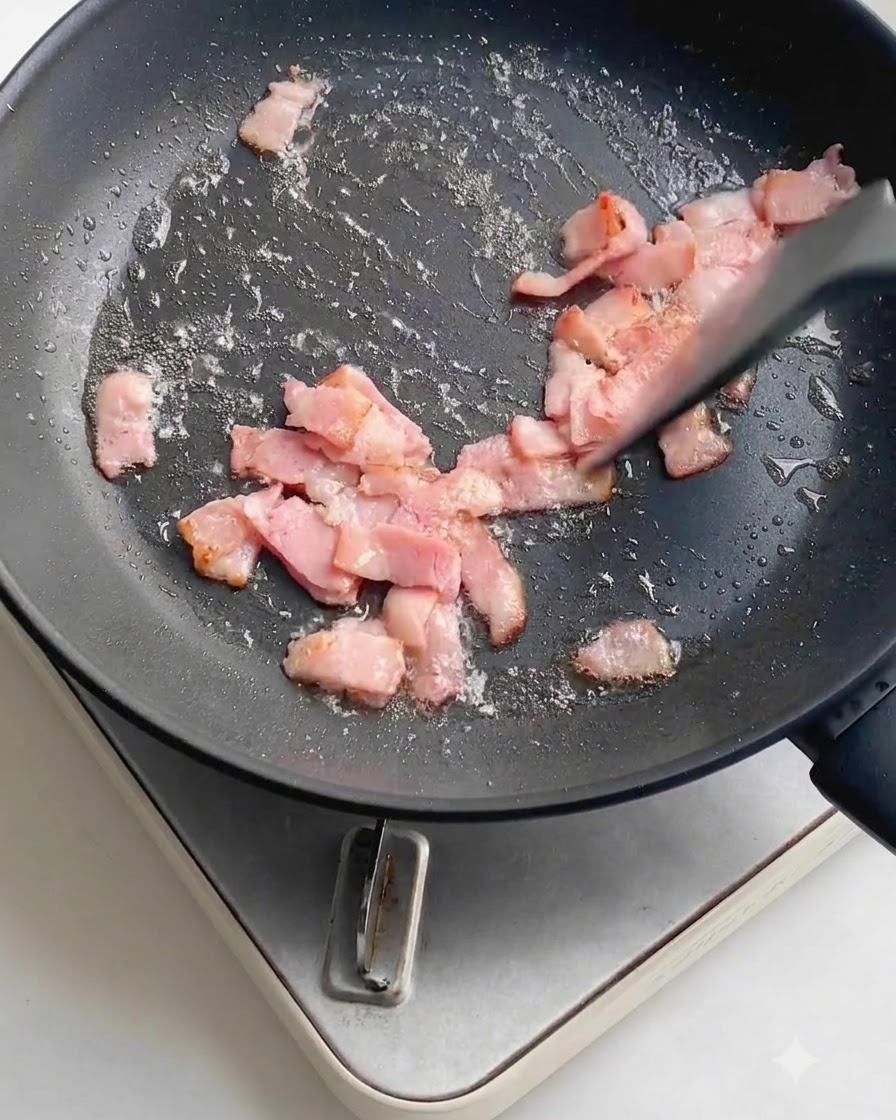 Fresh minced garlic scattered over the frying bacon pieces inside a dark non-stick frying pan.