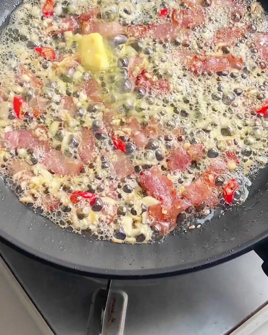 Cooked pasta noodles being added to a frying pan filled with bubbling bacon, melted butter, minced garlic, and sliced red chilies.