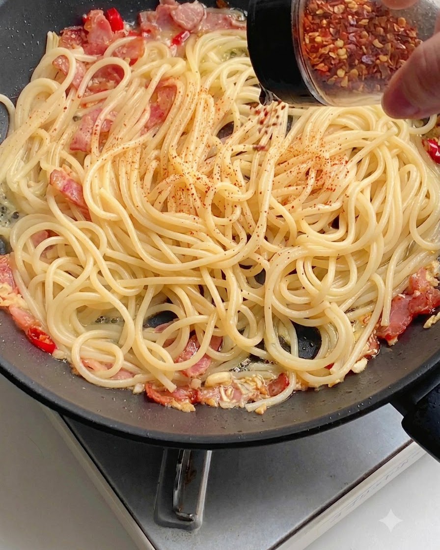 Red chili powder being sprinkled evenly over spaghetti in a pan.