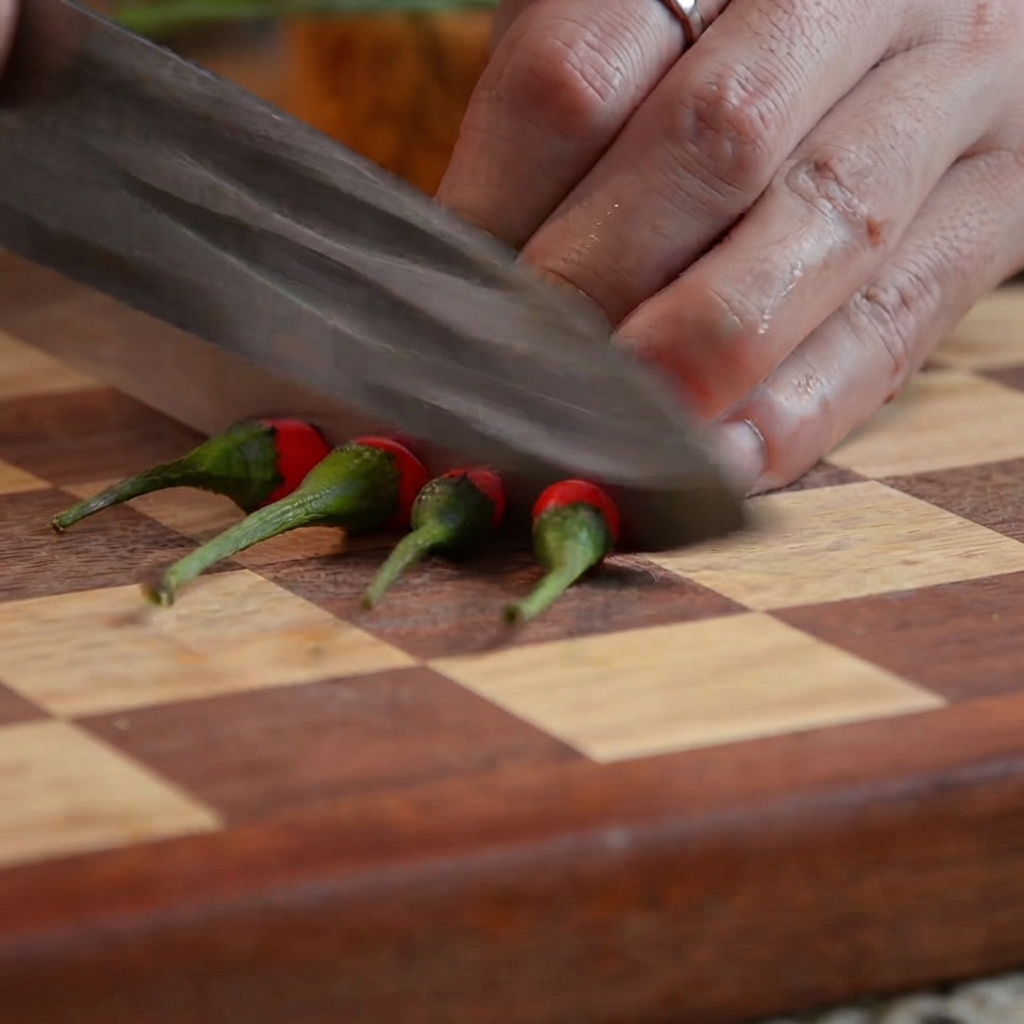 A kitchen knife cutting red chili peppers into small segments on a wooden cutting board.