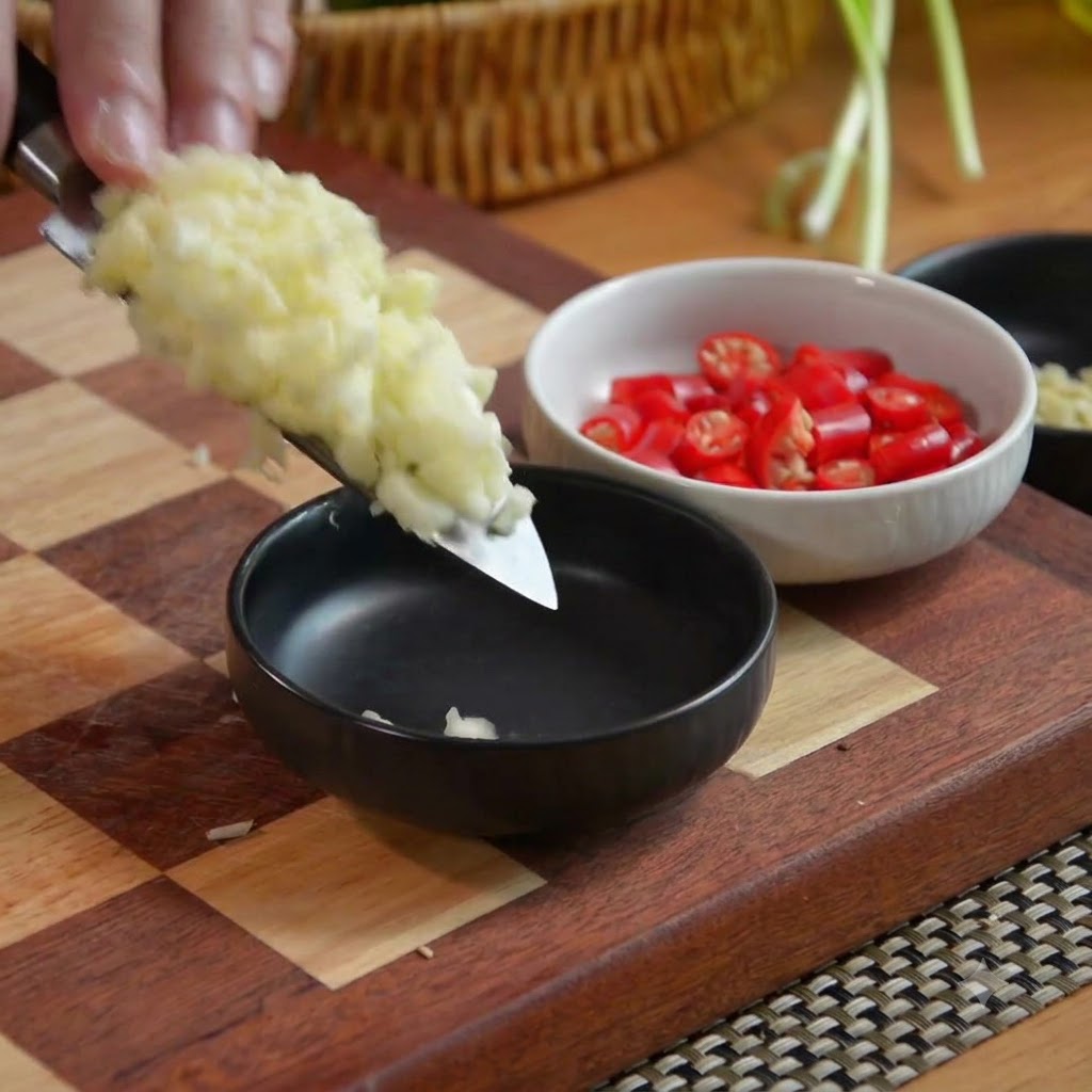 Minced garlic on a knife blade being transferred into a black bowl alongside a bowl of chopped red chilies.