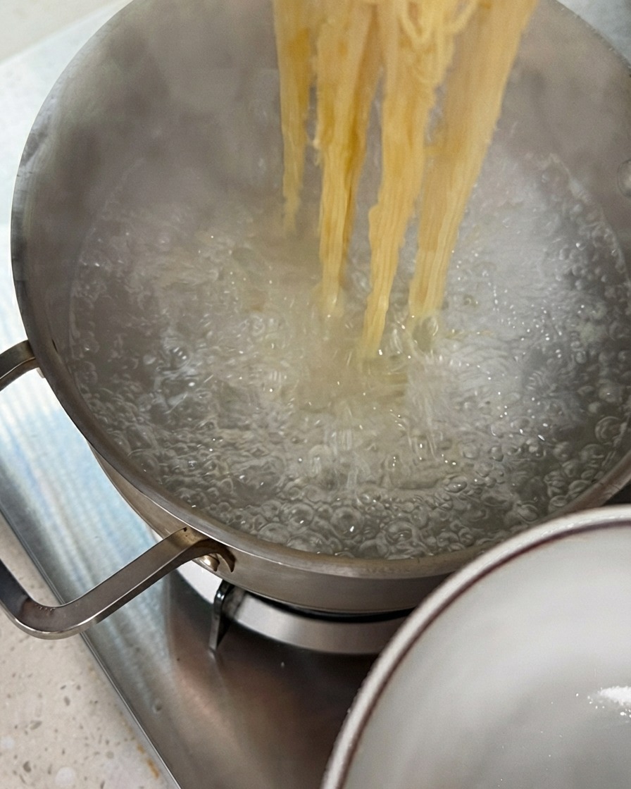 Tongs lifting strands of cooked noodles out of a boiling pot of water.