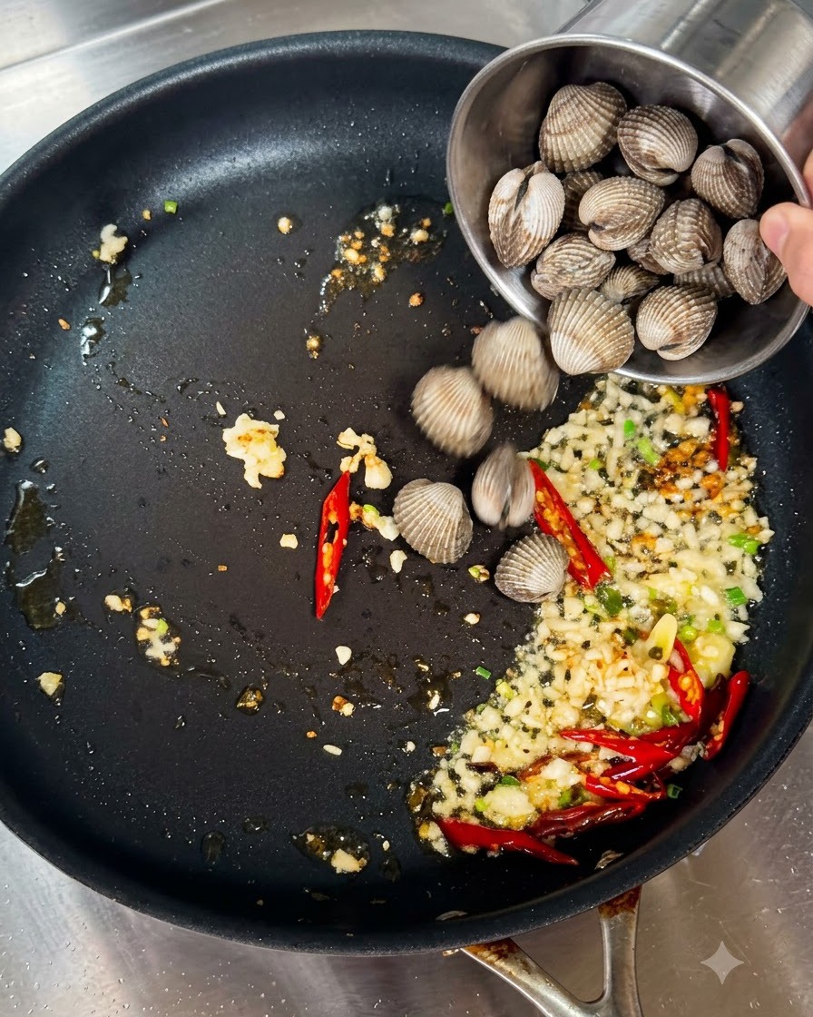Fresh clams being poured from a small metal cup into a frying pan with sautéed garlic and chilies.
