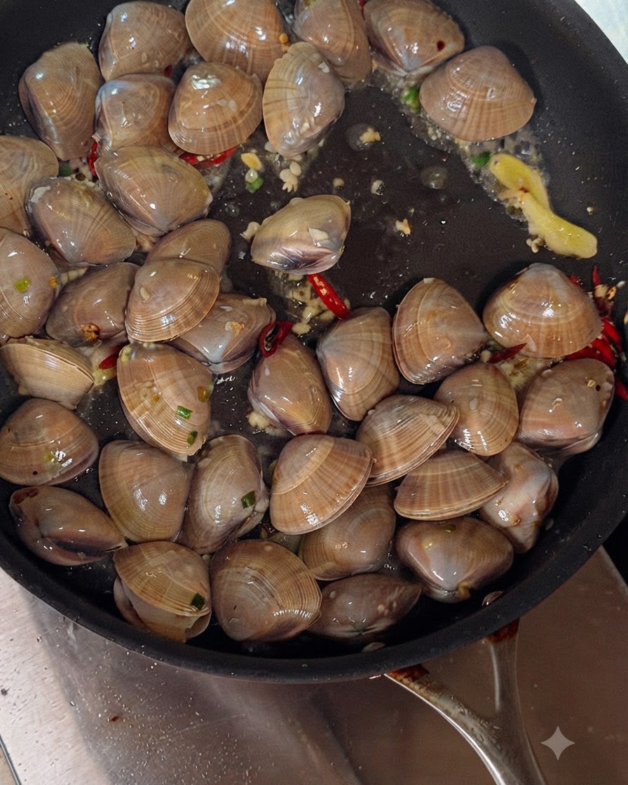 White wine being poured from a bottle into a sizzling pan filled with clams, garlic, and chili peppers.