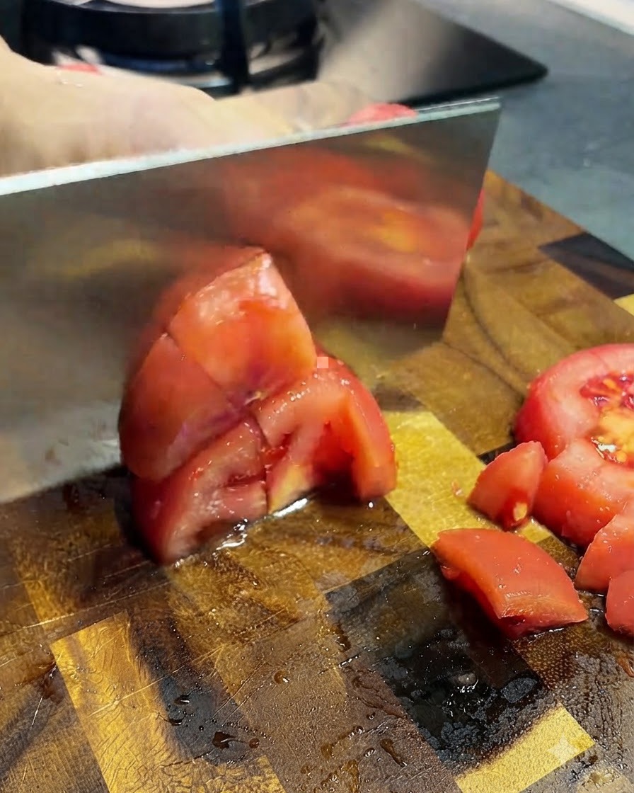 Peeled tomatoes being sliced into small pieces with a cleaver on a wooden board.