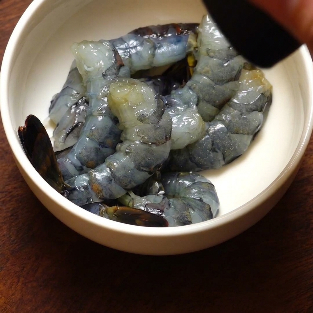 A bowl of peeled black tiger shrimp being seasoned with freshly ground black pepper from a grinder.