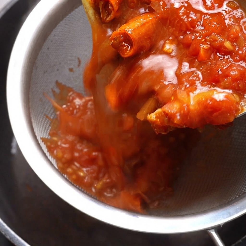 Cooked tomato sauce being poured through a metal sieve with a spatula pressing down the contents.