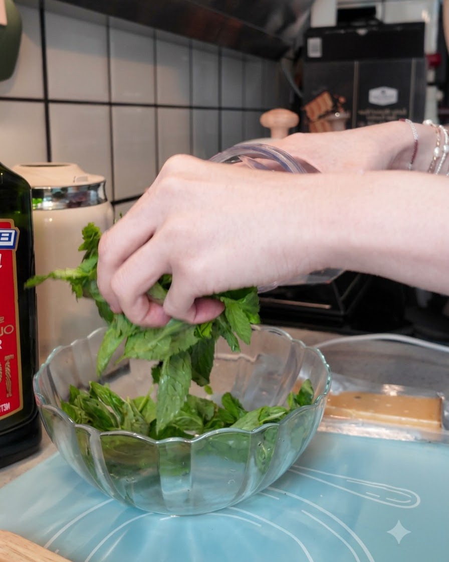 Fresh basil leaves being loaded into a clear blender cup alongside garlic and pine nuts.