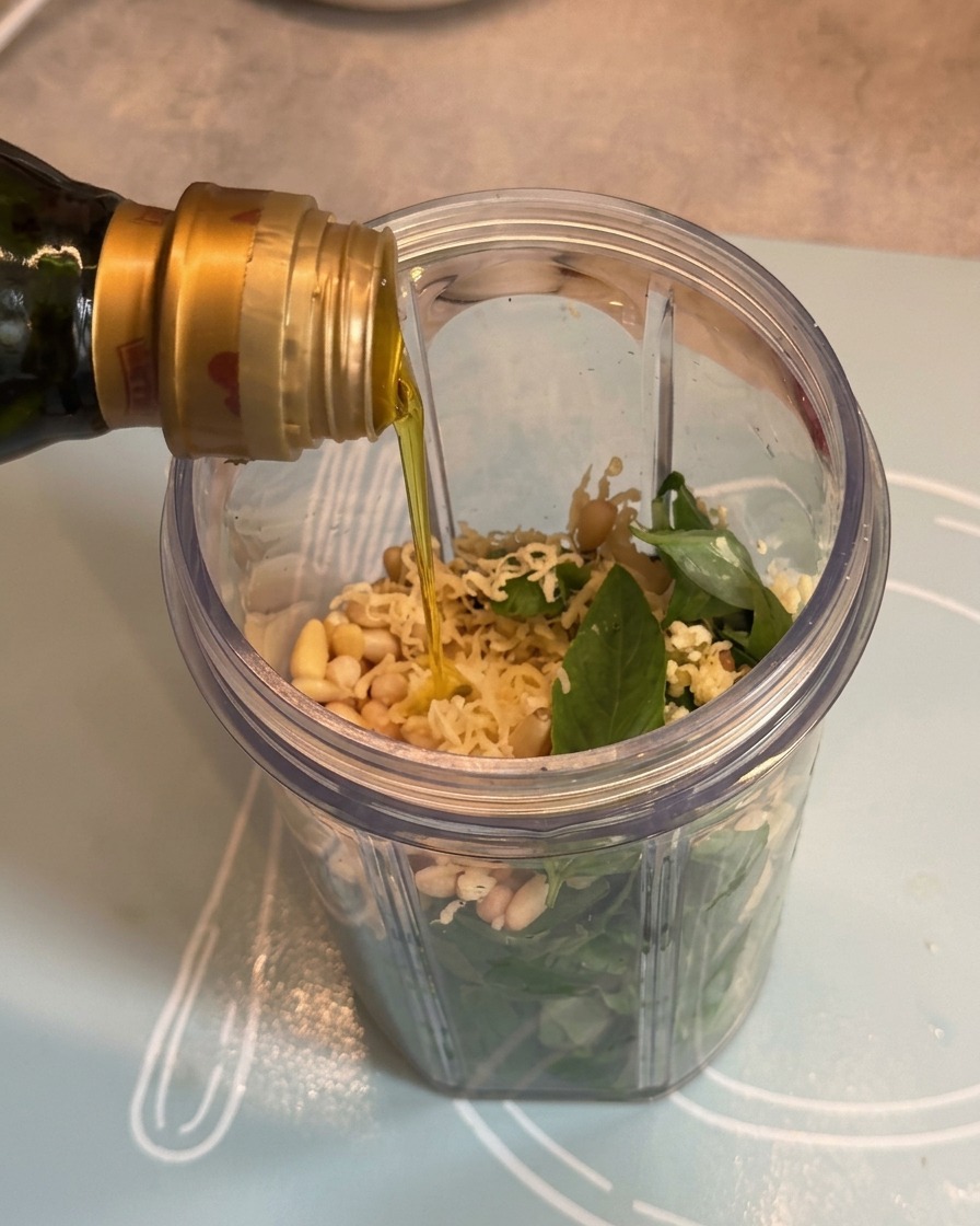 Olive oil being poured from a bottle into the blender cup over the pesto ingredients.