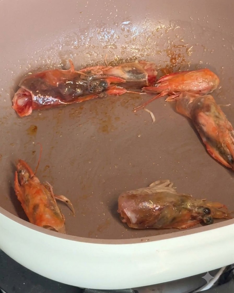 Peeled shrimp and shrimp heads sizzling in a pan while being seasoned with spices.