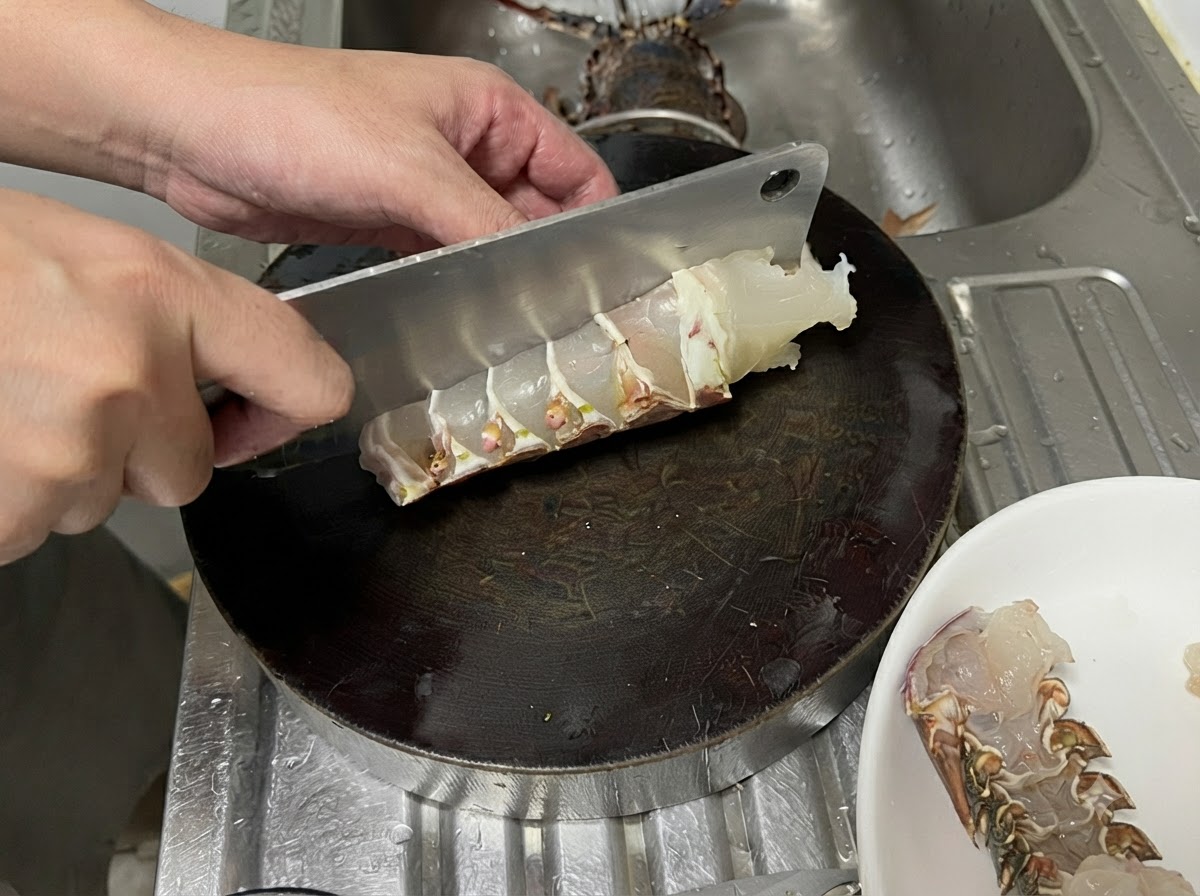 A large metal cleaver slicing through a raw lobster tail on a dark round cutting board to create thick, even pieces.