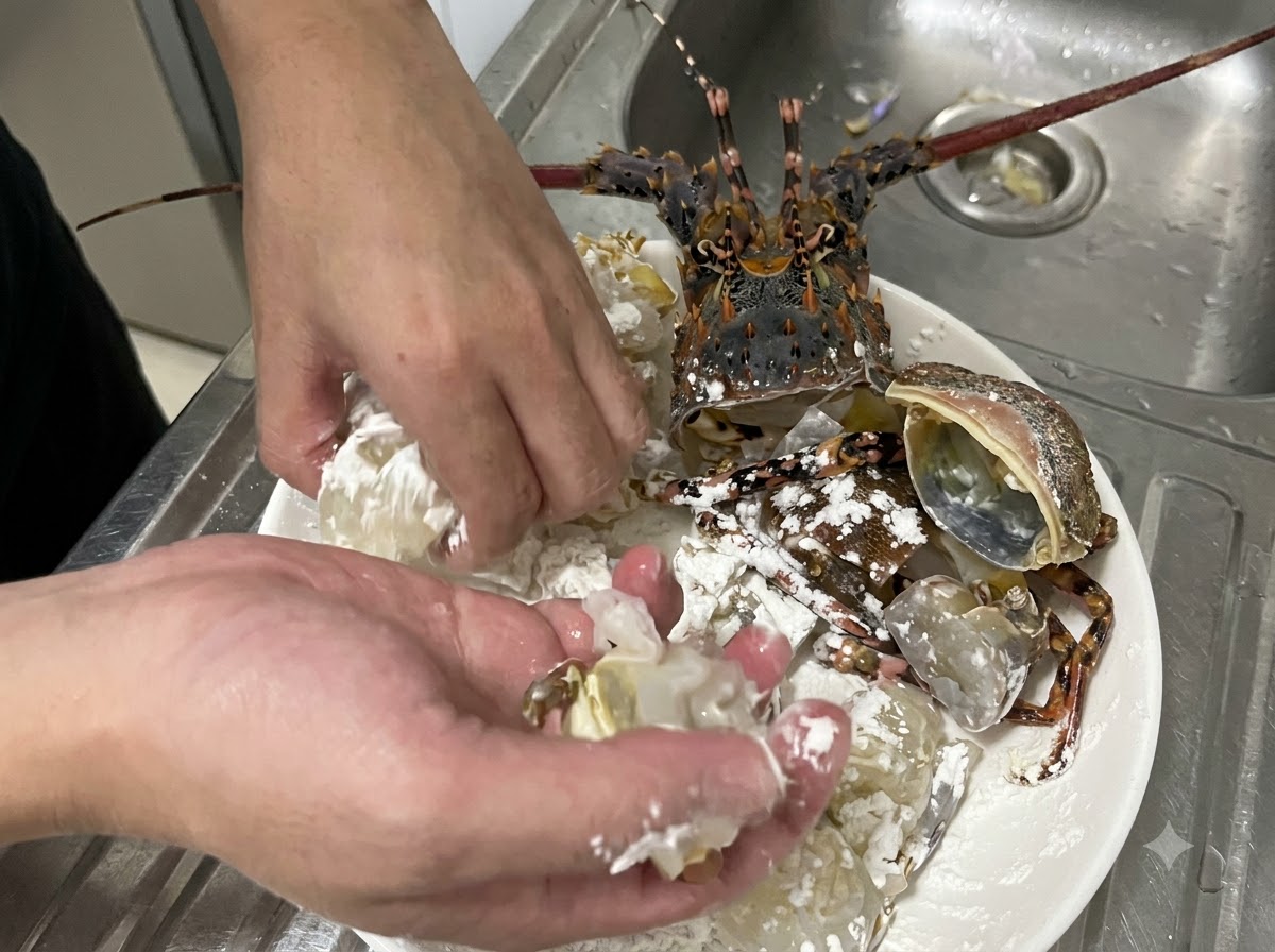 Hands actively tossing chunks of raw lobster meat and shell in white cornstarch on a plate to ensure an even coating.