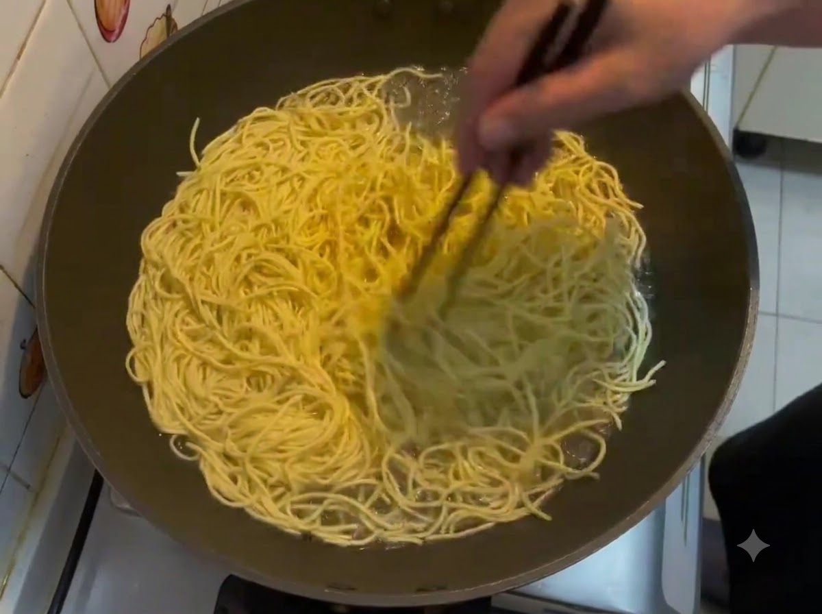 Chopsticks stirring a large batch of yellow E-fu noodles boiling in a wok.