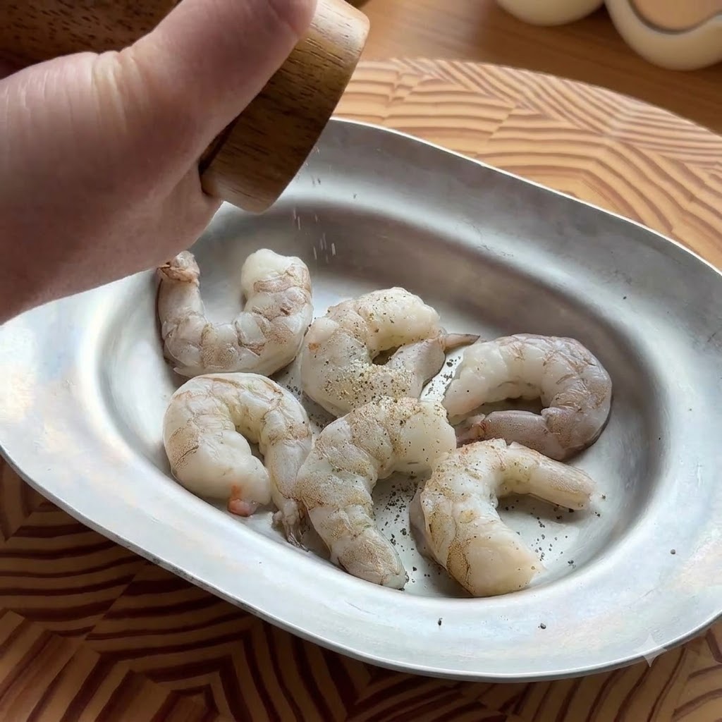 A hand using a wooden pepper grinder to season raw, peeled shrimp in an oval metal tray.