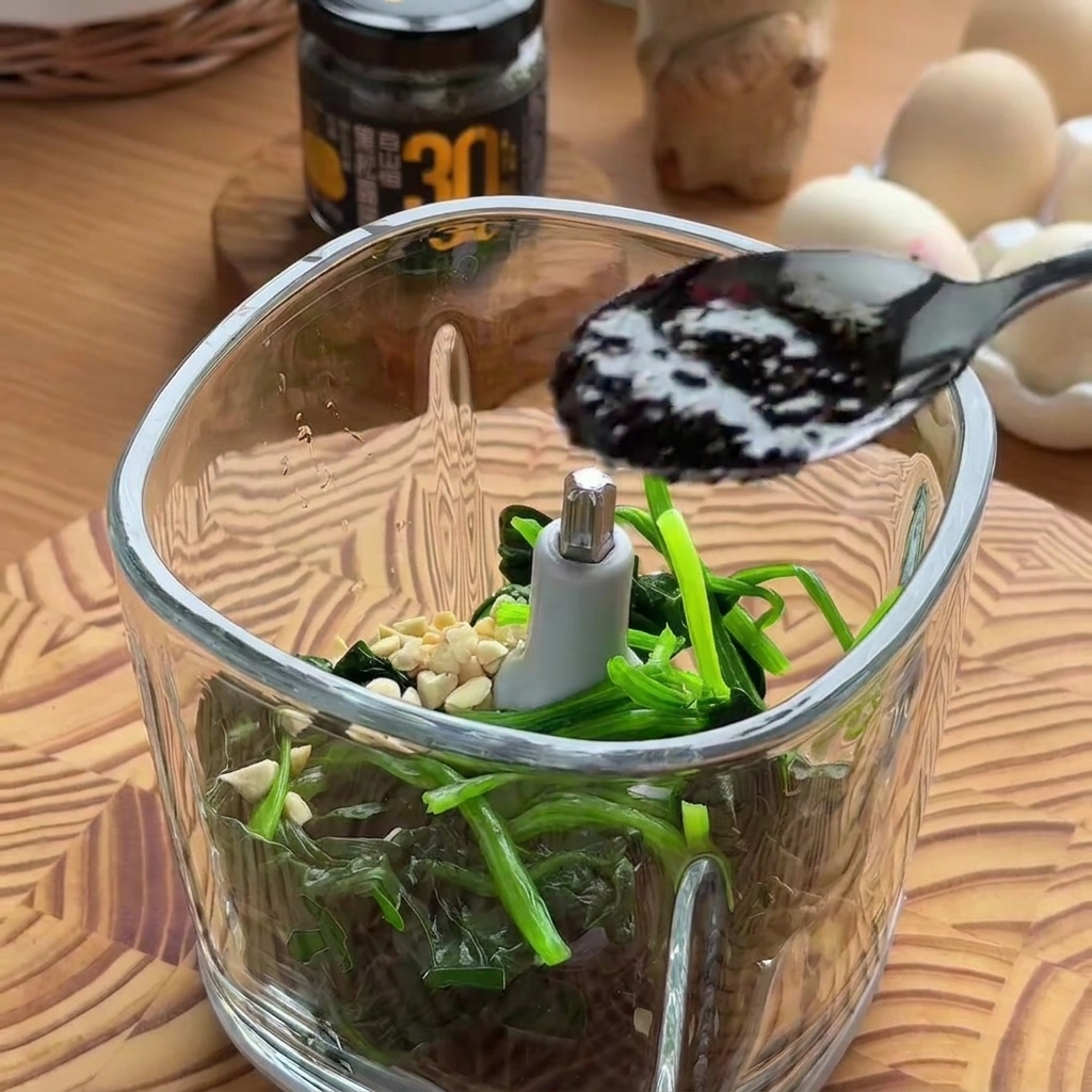 A black spoon dropping dark, rich black truffle sauce into a glass food processor bowl filled with spinach and nuts.