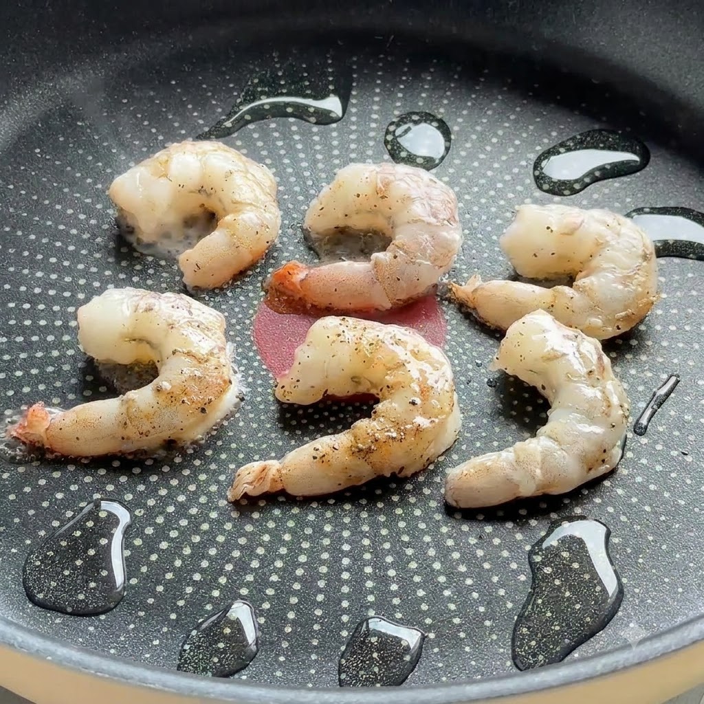 Seasoned shrimp sizzling in a non-stick frying pan with drops of oil.