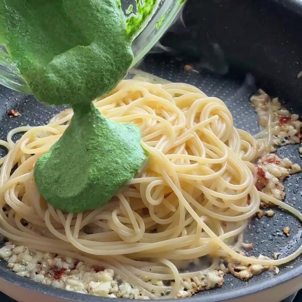 Green pesto sauce being poured from a glass bowl onto spaghetti noodles in a frying pan.