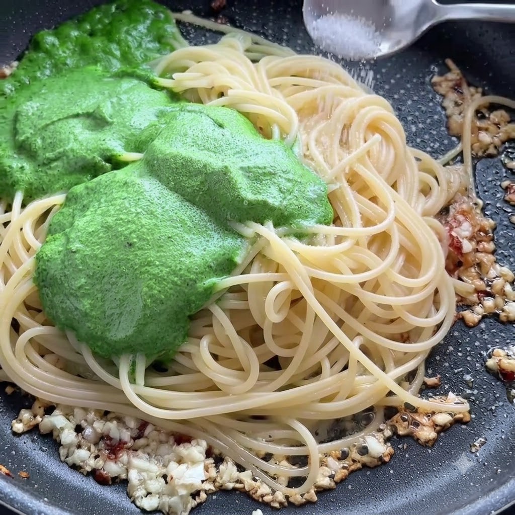A spoonful of salt being sprinkled over spaghetti noodles and green pesto sauce in a skillet.