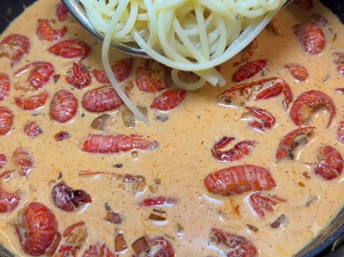 Tongs lifting boiled spaghetti noodles into a sizzling pan filled with creamy pink sauce and red crayfish tails.