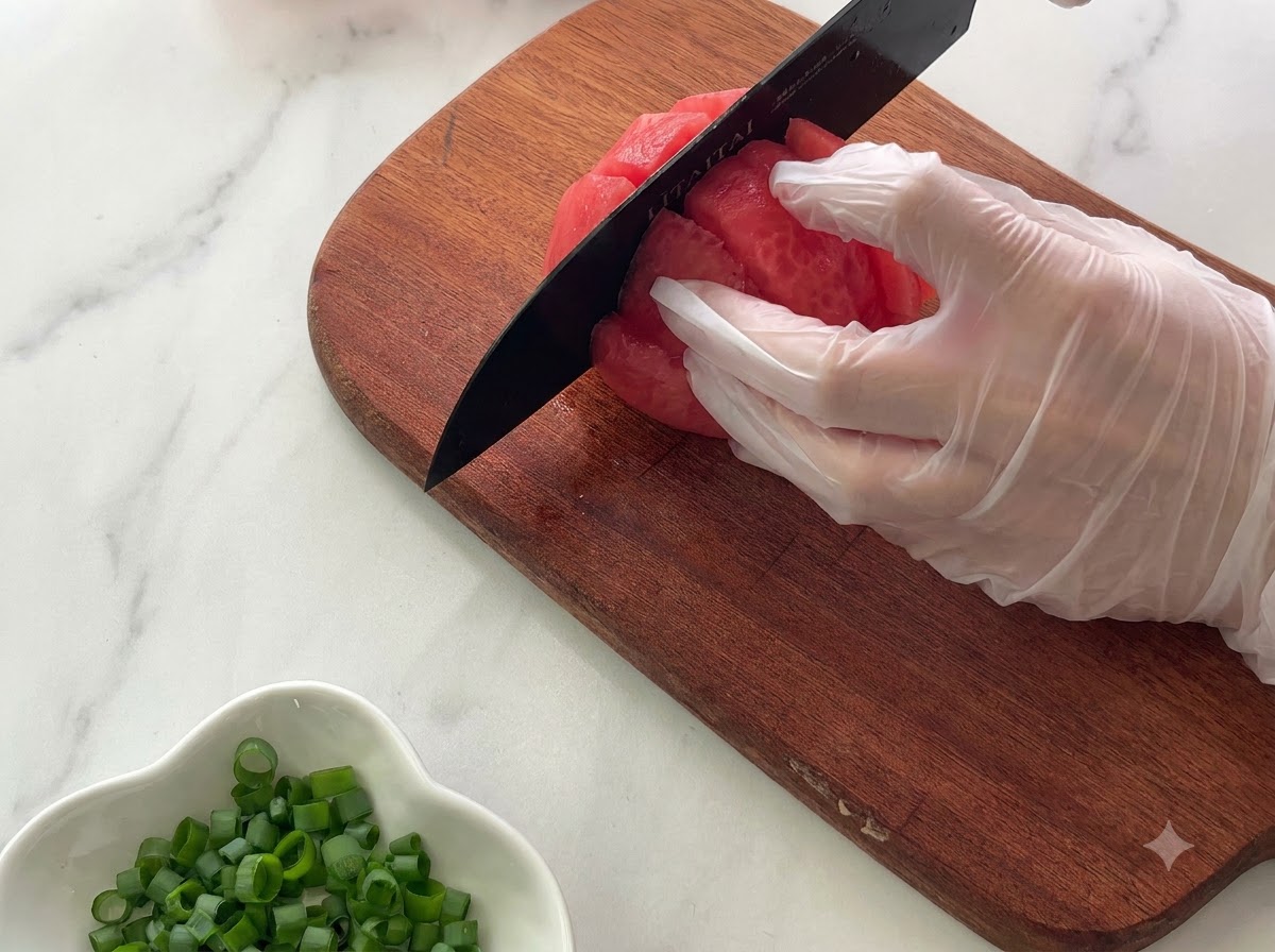 A gloved hand holding a tomato on a wooden cutting board while slicing it into chunks with a black knife.