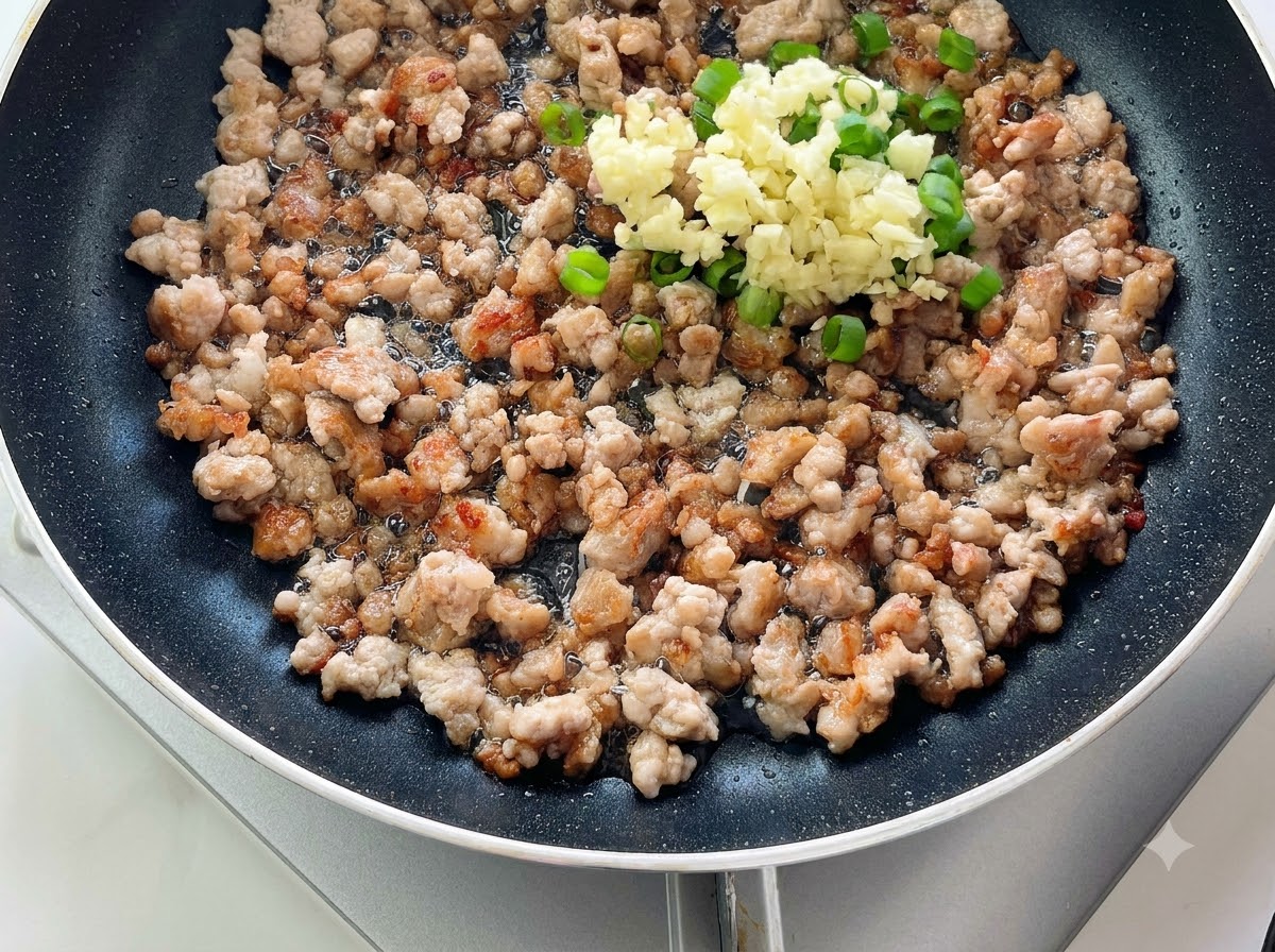 Chopped garlic and green onions piled on top of cooked minced meat in a pan.