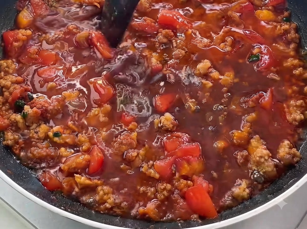 A close-up of dark liquid sauce being poured into a pan filled with browned minced meat and diced tomatoes.