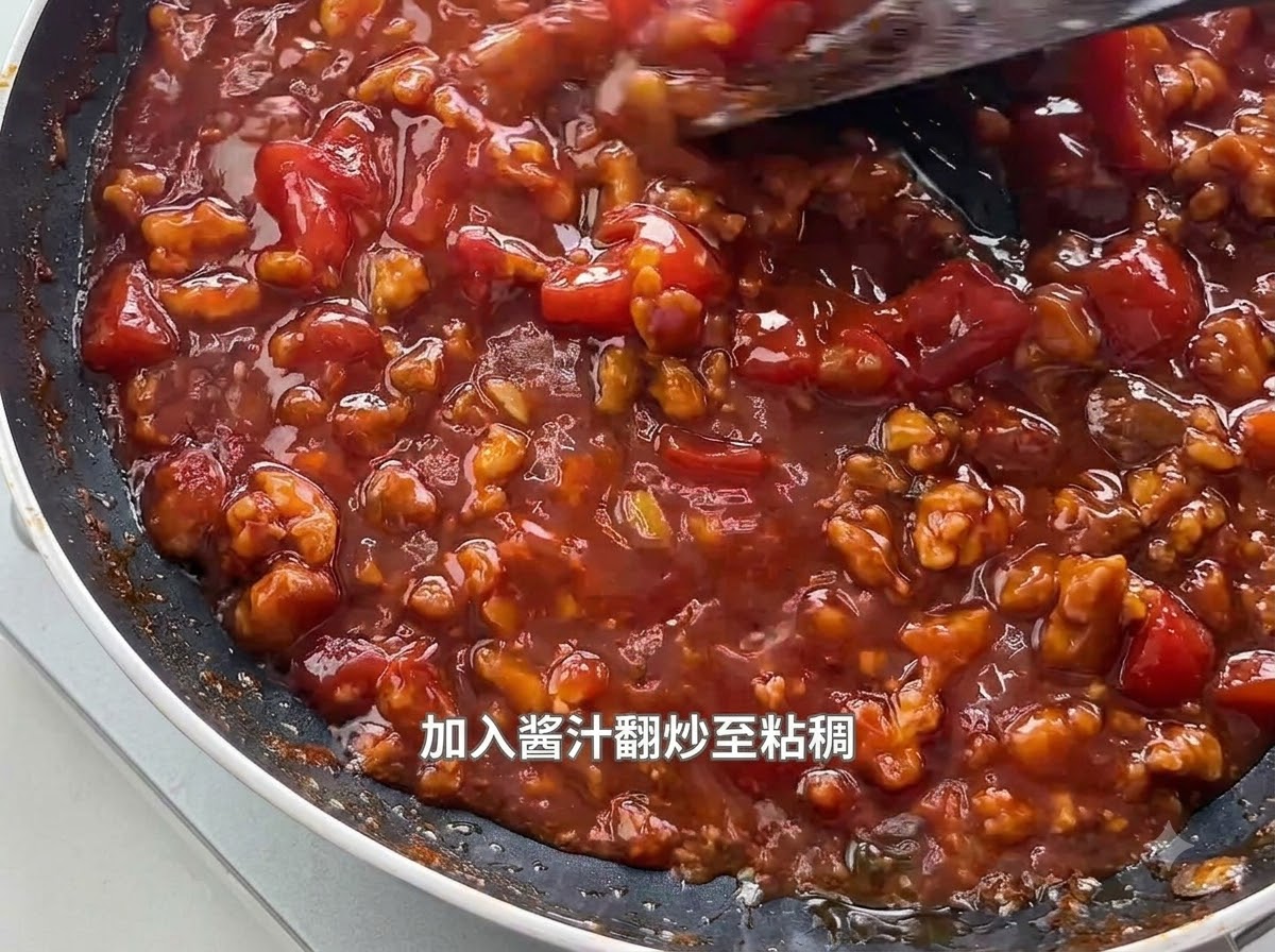 A spatula stirring a thick, bubbling tomato and minced meat sauce simmering in a black frying pan.