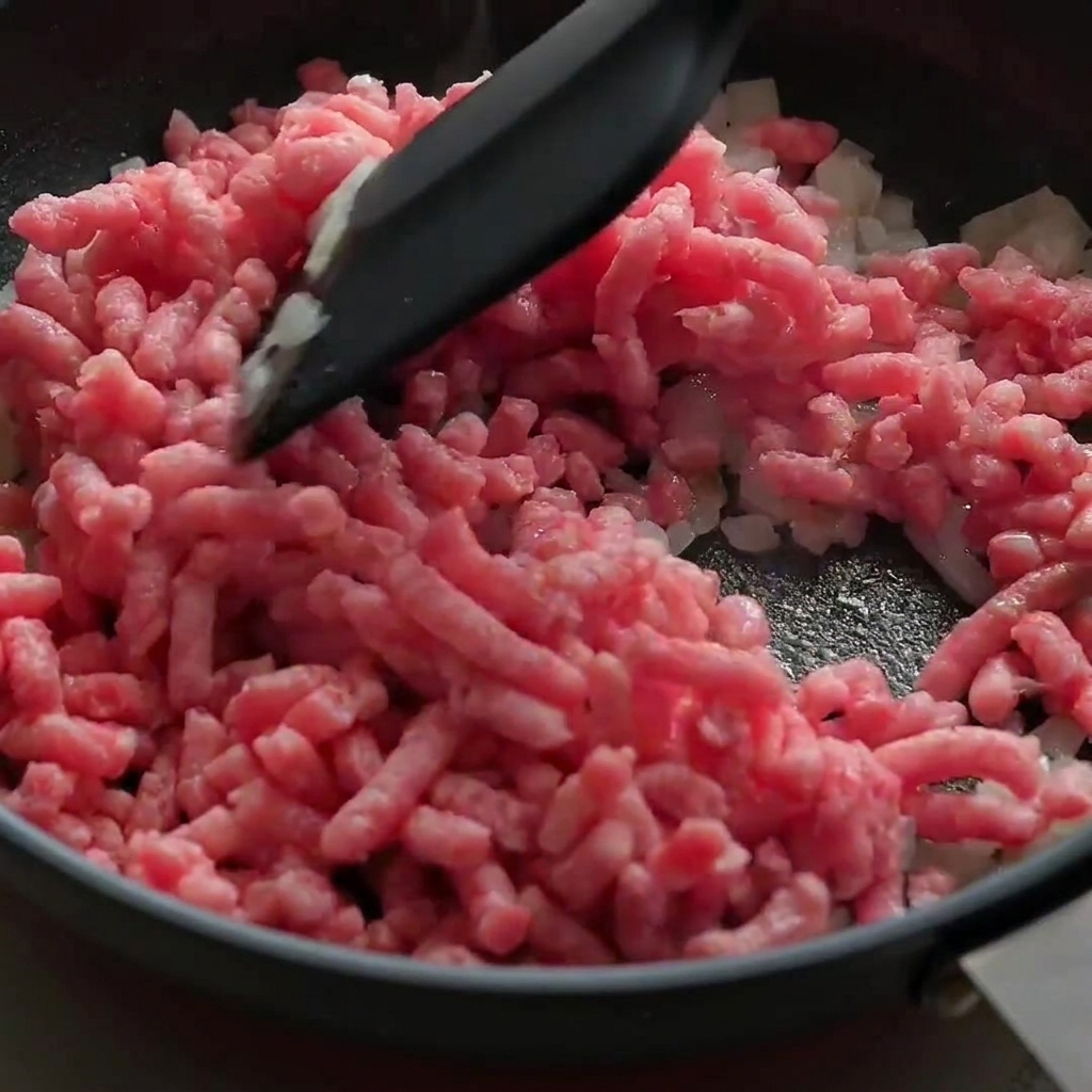 Raw pink minced beef being broken apart and stir-fried with onions in a black skillet.
