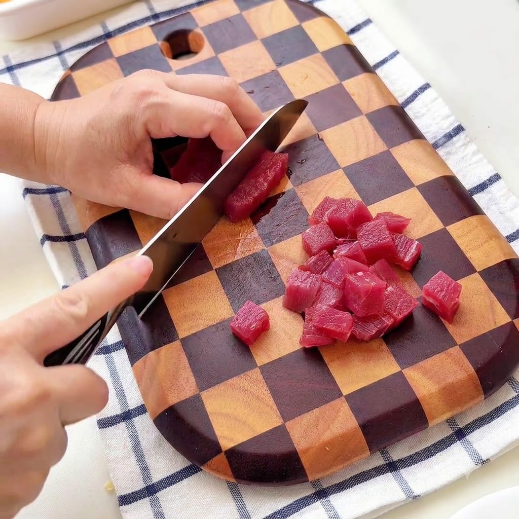 A person cutting beef into small cubes on a checkered wooden cutting board.