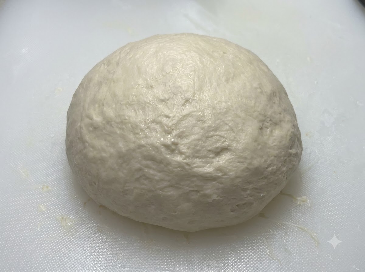 A smooth rounded dough ball resting on a white board prior to being covered.
