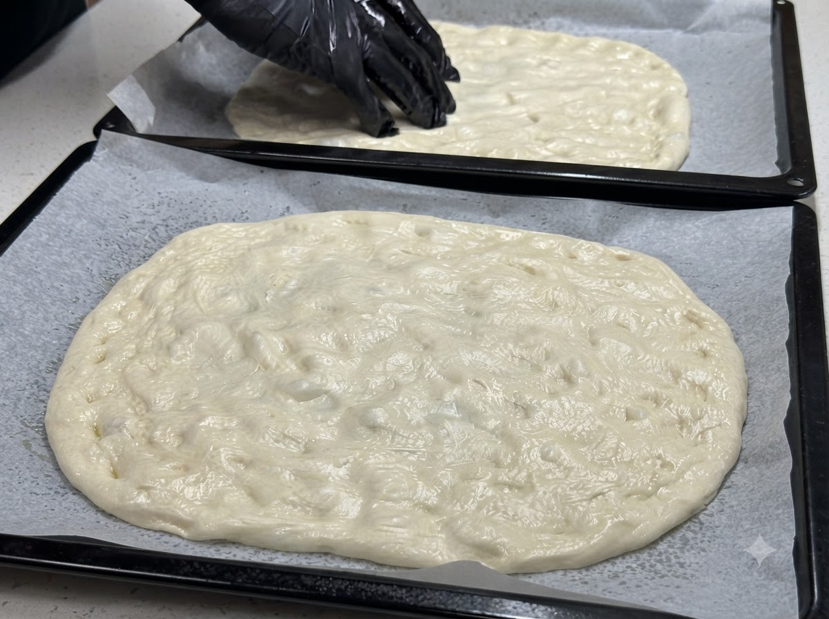Two baking sheets with flattened dough, with a gloved hand placing cherry tomato halves across the surface.