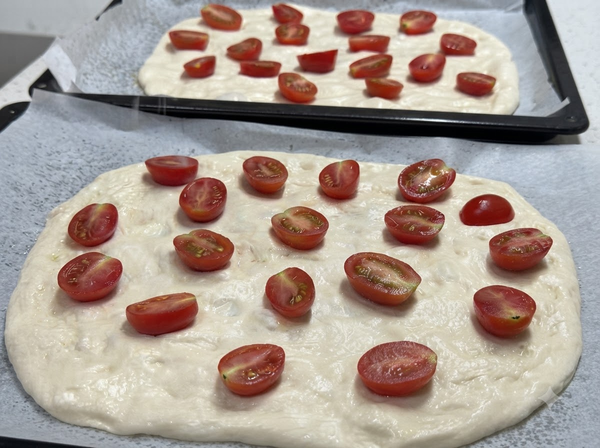 A close-up of two stretched pizza dough bases topped with halved cherry tomatoes, ready for seasoning.