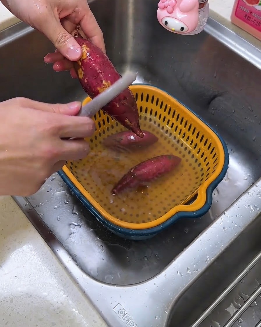 A person peeling raw sweet potatoes over a kitchen sink using a vegetable peeler.