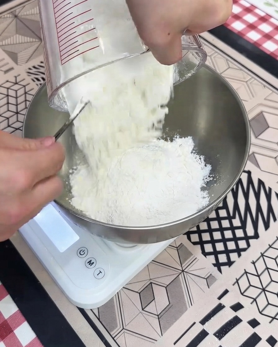 Dry high-protein flour being poured from a measuring cup into a metal bowl sitting on a white digital scale.