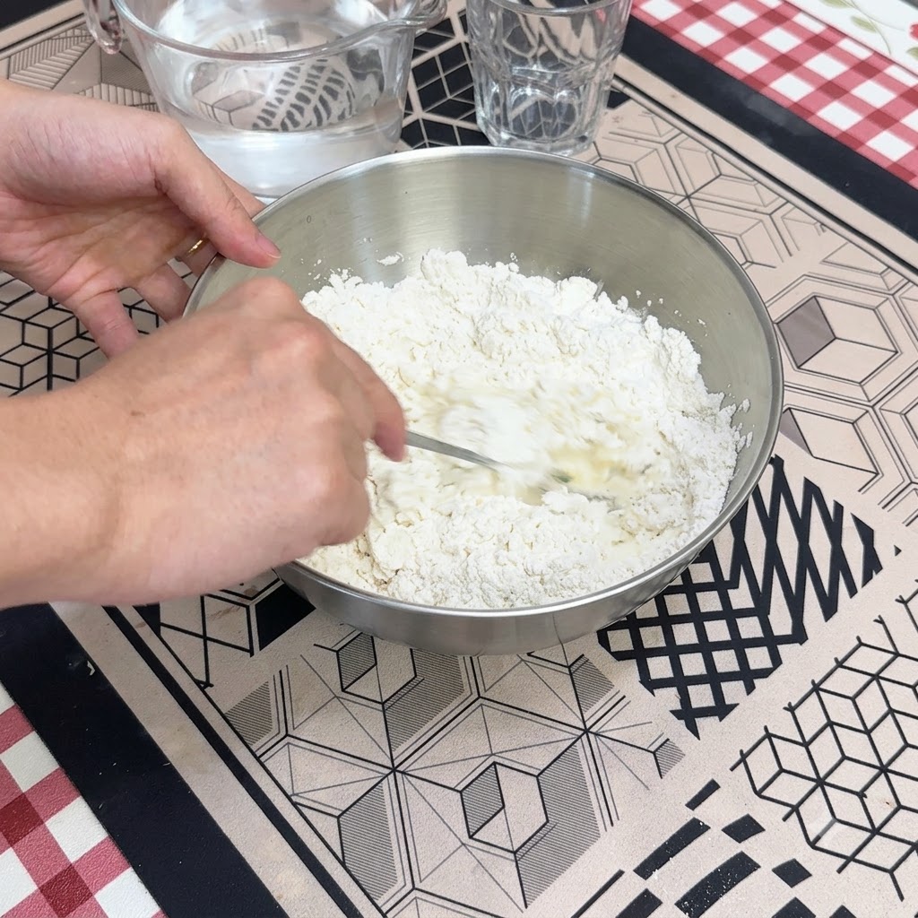 A hand using a spoon to stir water into dry flour inside a metal mixing bowl.