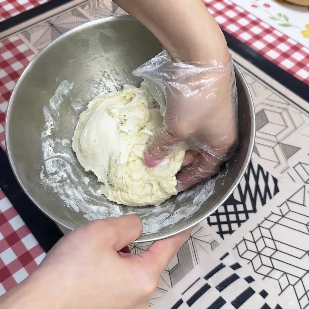 A gloved hand kneading wet pizza dough inside a stainless steel mixing bowl.